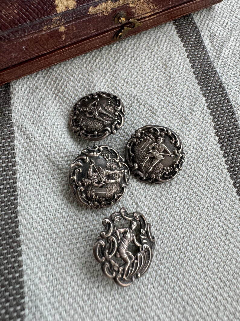 May include: Four silver-toned buttons with ornate designs featuring figures in various poses. The buttons have a raised, textured surface and intricate details.