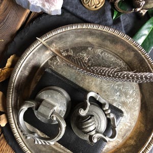 Peut inclure: Deux poignées de tiroir en argent antique avec des motifs ornés, l'une avec un bouton rond et l'autre avec un bouton cannelé, sont présentées sur un livre relié en cuir noir sur un plateau en argent.