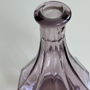 May include: A close-up of a lavender-colored glass bottle. The bottle has a geometric, faceted design and a rounded opening. The glass is transparent, allowing light to pass through, and the bottle's shape is elegant and decorative.