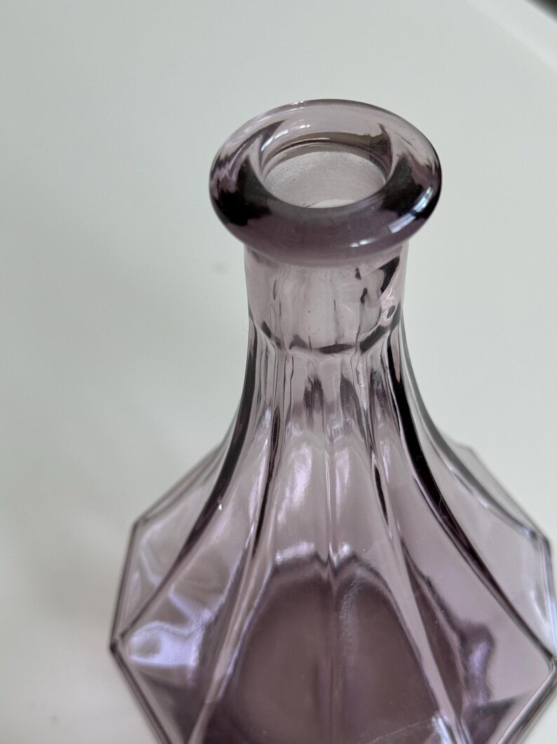 May include: A close-up of a lavender-colored glass bottle. The bottle has a geometric, faceted design and a rounded opening. The glass is transparent, allowing light to pass through, and the bottle's shape is elegant and decorative.