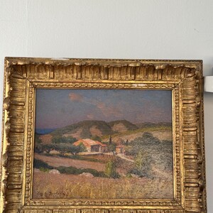 Puede incluir: Una pintura al &oacute;leo enmarcada que representa una escena de paisaje con un peque&ntilde;o pueblo enclavado en las colinas. La pintura presenta una paleta de colores c&aacute;lidos con tonos de marr&oacute;n, verde y azul. El marco es ornamentado y de color dorado.
