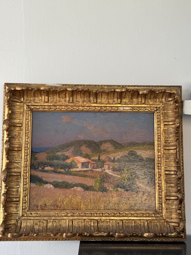Puede incluir: Una pintura al &oacute;leo enmarcada que representa una escena de paisaje con un peque&ntilde;o pueblo enclavado en las colinas. La pintura presenta una paleta de colores c&aacute;lidos con tonos de marr&oacute;n, verde y azul. El marco es ornamentado y de color dorado.
