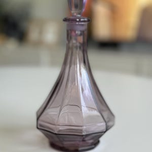 May include: A vintage, amethyst-colored glass perfume bottle with a faceted, geometric design. The bottle has a matching stopper and a flared base. The bottle is approximately 6 inches tall.