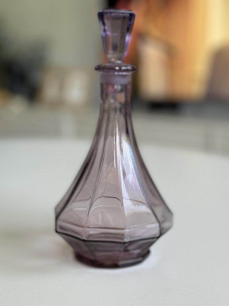 May include: A vintage, amethyst-colored glass perfume bottle with a faceted, geometric design. The bottle has a matching stopper and a flared base. The bottle is approximately 6 inches tall.