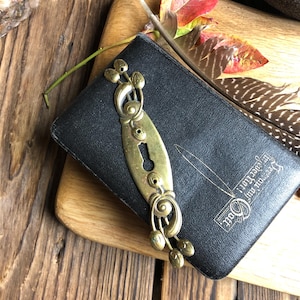 Op de afbeelding: Een zwart lederen gebonden dagboek met een messing sluiting met een sleutelgat en een bloemmotief. Het dagboek heeft de tekst "In God we trust" en "May you find your way" op de omslag.