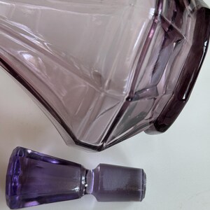 May include: A vintage, amethyst-colored glass perfume bottle with a geometric design. The bottle has a faceted shape and is accompanied by a matching stopper. The glass has a translucent quality, allowing light to pass through.