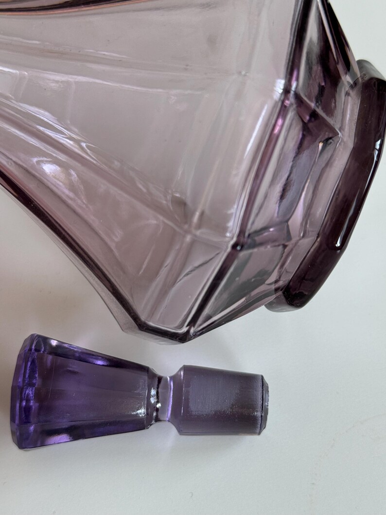 May include: A vintage, amethyst-colored glass perfume bottle with a geometric design. The bottle has a faceted shape and is accompanied by a matching stopper. The glass has a translucent quality, allowing light to pass through.