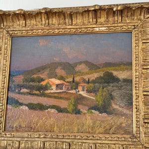 Puede incluir: Una pintura al &oacute;leo enmarcada que representa un paisaje rural con un peque&ntilde;o pueblo enclavado entre colinas onduladas. La pintura presenta una paleta de colores c&aacute;lidos con tonos de marr&oacute;n, verde y azul. El cielo es de un azul suave con toques de nubes rosadas y naranjas. El pueblo est&aacute; formado por unas pocas casas sencillas con techos de tejas rojas. Las colinas est&aacute;n cubiertas de vegetaci&oacute;n exuberante, y hay &aacute;rboles dispersos por toda la escena. La pintura est&aacute; enmarcada en un marco dorado con detalles ornamentados.