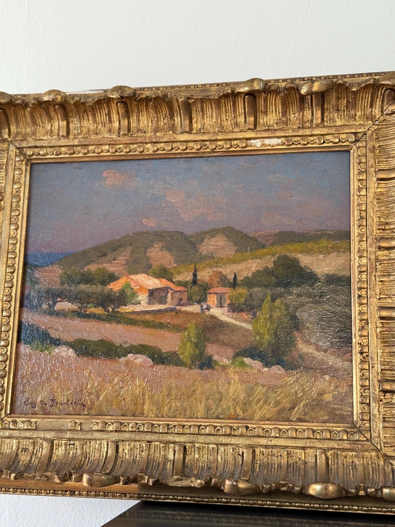 Puede incluir: Una pintura al &oacute;leo enmarcada que representa un paisaje rural con un peque&ntilde;o pueblo enclavado entre colinas onduladas. La pintura presenta una paleta de colores c&aacute;lidos con tonos de marr&oacute;n, verde y azul. El cielo es de un azul suave con toques de nubes rosadas y naranjas. El pueblo est&aacute; formado por unas pocas casas sencillas con techos de tejas rojas. Las colinas est&aacute;n cubiertas de vegetaci&oacute;n exuberante, y hay &aacute;rboles dispersos por toda la escena. La pintura est&aacute; enmarcada en un marco dorado con detalles ornamentados.