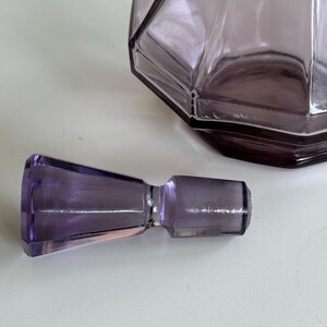 May include: A faceted, amethyst-colored glass bottle with a matching stopper. The bottle has a geometric design and is partially visible. The stopper is cylindrical with a flared end. The items are on a white surface.