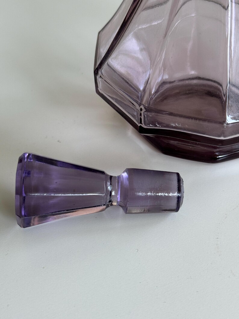 May include: A faceted, amethyst-colored glass bottle with a matching stopper. The bottle has a geometric design and is partially visible. The stopper is cylindrical with a flared end. The items are on a white surface.
