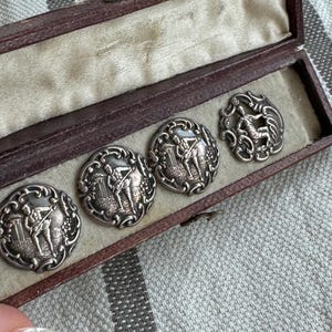 May include: A set of five silver-toned buttons in a brown velvet-lined box. Each button features a raised design of two figures playing a musical instrument.