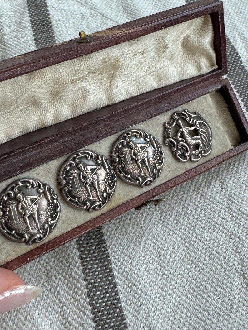 May include: A set of five silver-toned buttons in a brown velvet-lined box. Each button features a raised design of two figures playing a musical instrument.