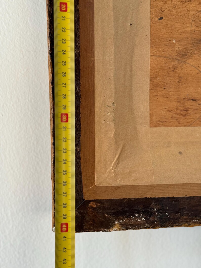 Puede incluir: Primer plano del borde inferior de un marco de imagen de madera con un respaldo de papel marr&oacute;n. Una cinta m&eacute;trica amarilla es visible a lo largo del borde izquierdo del marco, con los n&uacute;meros del 20 al 43 visibles.