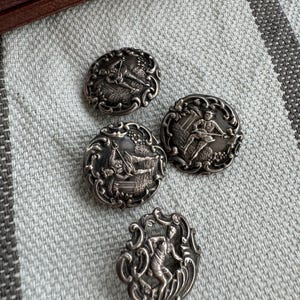 May include: Four silver-toned buttons with intricate designs featuring figures in various poses. The buttons are round with a raised, detailed design.