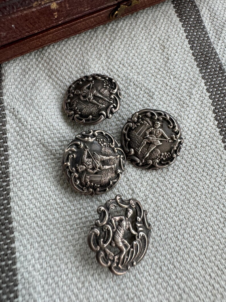 May include: Four silver-toned buttons with intricate designs featuring figures in various poses. The buttons are round with a raised, detailed design.
