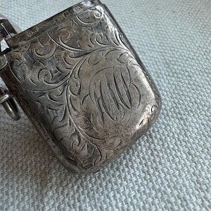 May include: A silver match safe with an engraved floral design and the initials "H.A.W." on the front.