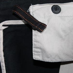 May include: A black garment featuring a white fabric pocket secured with a dark button. A black fabric strap with orange stitching is attached near the pocket. The garment includes white piping along the seams, creating a high-contrast design.