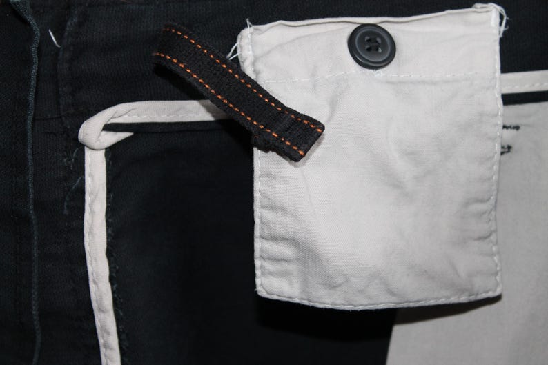 May include: A black garment featuring a white fabric pocket secured with a dark button. A black fabric strap with orange stitching is attached near the pocket. The garment includes white piping along the seams, creating a high-contrast design.