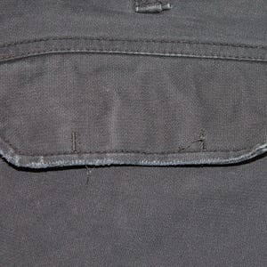 May include: A dark charcoal gray fabric pocket with a rectangular flap featuring stitched edges and visible signs of wear along the bottom seam. The pocket is integrated into a matching dark gray textile surface with a visible belt loop above.