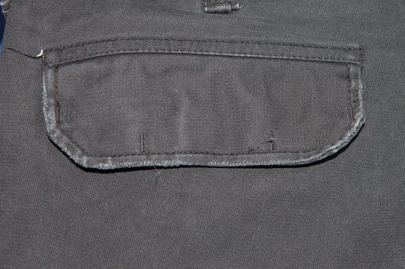 May include: A dark charcoal gray fabric pocket with a rectangular flap featuring stitched edges and visible signs of wear along the bottom seam. The pocket is integrated into a matching dark gray textile surface with a visible belt loop above.
