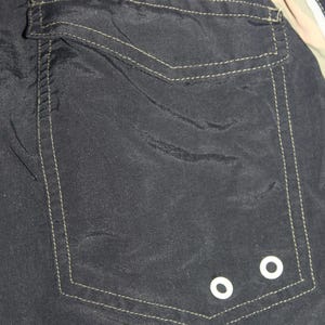 May include: A black fabric garment featuring a large rectangular patch pocket with a flap closure. The pocket is accented with visible light-colored stitching and two small white circular metal grommets located at the bottom right corner.