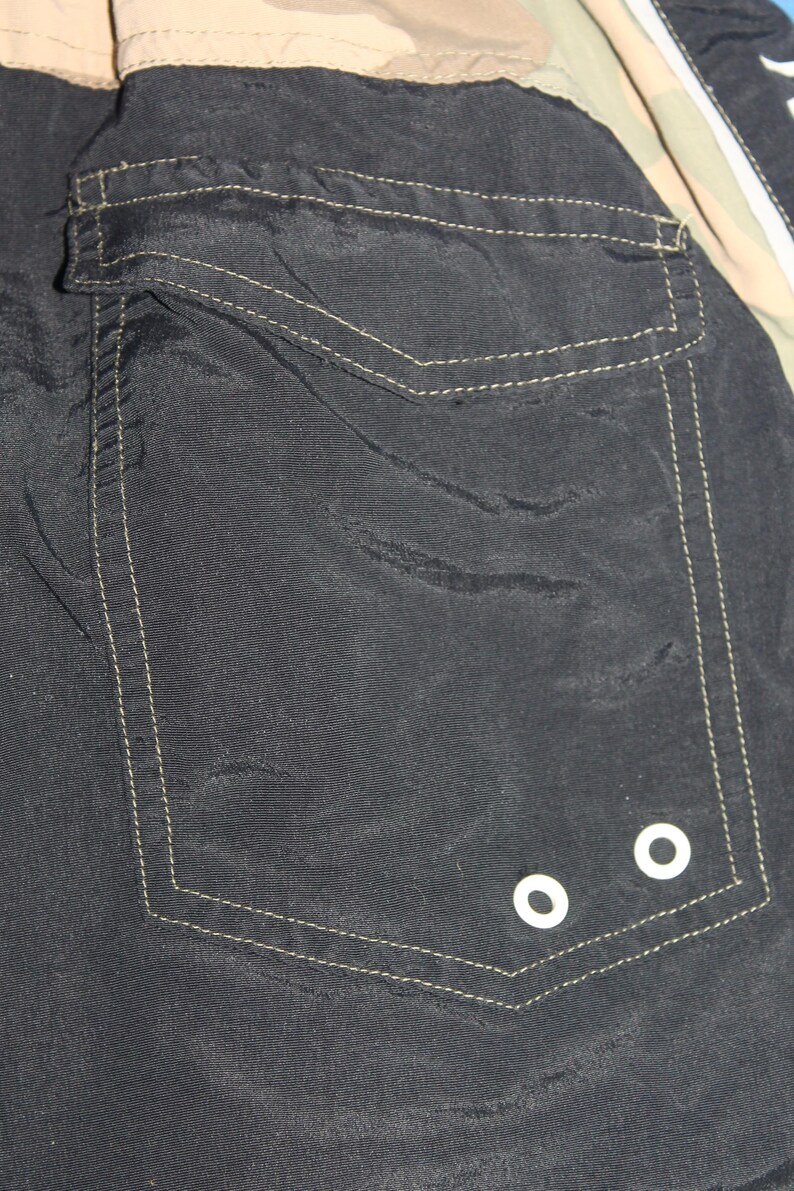 May include: A black fabric garment featuring a large rectangular patch pocket with a flap closure. The pocket is accented with visible light-colored stitching and two small white circular metal grommets located at the bottom right corner.