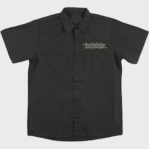 Può includere: Una camicia da lavoro nera a maniche corte con colletto, due tasche sul petto con chiusura a bottone e un'abbottonatura centrale. La parola incubus è stampata in un carattere bianco stilizzato con accenti verdi sopra la tasca sinistra.