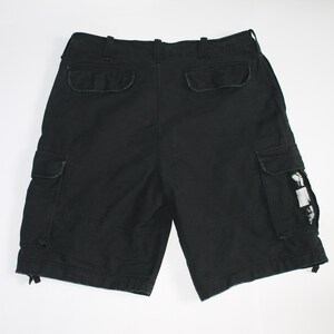 May include: Black cargo shorts featuring a waistband with belt loops, two back pockets with flaps, and two side cargo pockets. One side pocket displays a small white rectangular patch with a black graphic print. The leg openings have drawstrings.