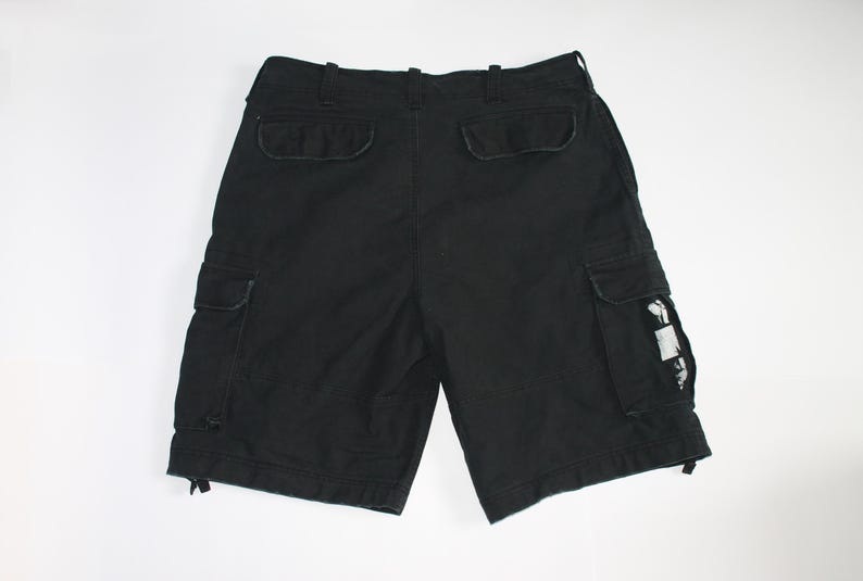 May include: Black cargo shorts featuring a waistband with belt loops, two back pockets with flaps, and two side cargo pockets. One side pocket displays a small white rectangular patch with a black graphic print. The leg openings have drawstrings.