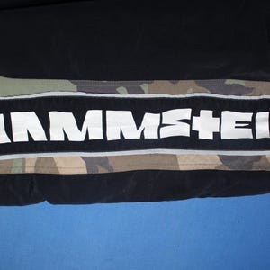 May include: Black and camouflage patterned pants featuring a wide horizontal black stripe down the side. The stripe contains the white text RAMMSTEIN in a bold, stylized font, bordered by thin reflective silver piping on the top and bottom edges.