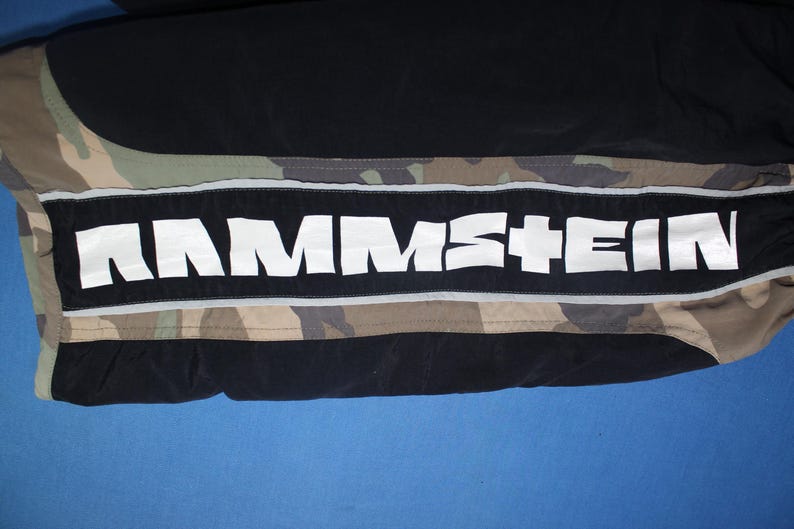 May include: Black and camouflage patterned pants featuring a wide horizontal black stripe down the side. The stripe contains the white text RAMMSTEIN in a bold, stylized font, bordered by thin reflective silver piping on the top and bottom edges.