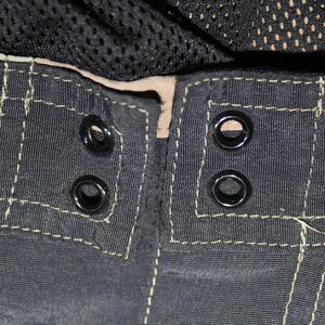 May include: A pair of black shorts featuring a waistband with four black metal eyelets arranged in two pairs. The fabric is accented with visible light-colored stitching along the seams and a tan inner lining, with black mesh fabric visible above.