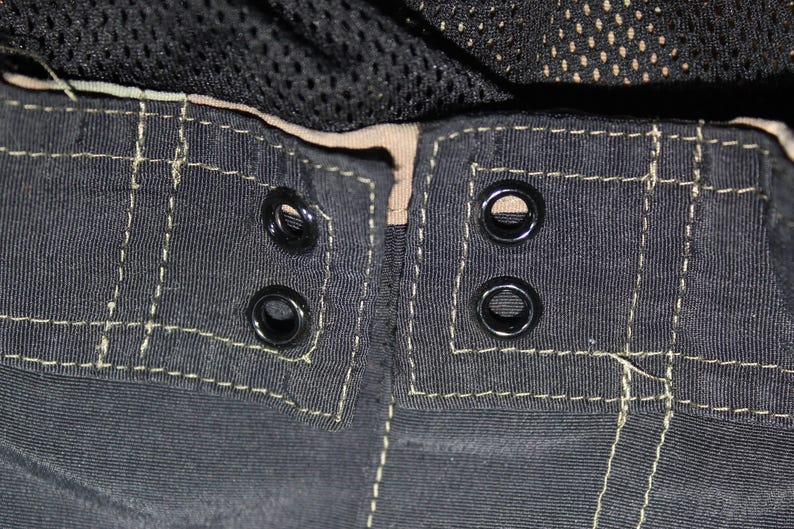 May include: A pair of black shorts featuring a waistband with four black metal eyelets arranged in two pairs. The fabric is accented with visible light-colored stitching along the seams and a tan inner lining, with black mesh fabric visible above.
