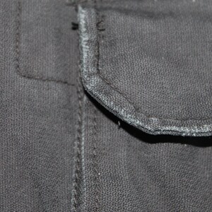 May include: A close-up view of a dark charcoal gray fabric garment featuring a rectangular pocket flap with visible stitching along the edges. The material has a textured, woven appearance with slight fraying along the seam of the pocket flap.