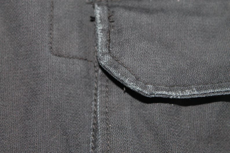 May include: A close-up view of a dark charcoal gray fabric garment featuring a rectangular pocket flap with visible stitching along the edges. The material has a textured, woven appearance with slight fraying along the seam of the pocket flap.