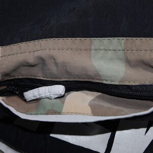 May include: A black garment featuring a horizontal zippered pocket with a camouflage-patterned interior lining. The pocket opening is framed by a light gray reflective trim, and a small white fabric pull tab is attached to the black zipper slider.