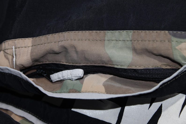 May include: A black garment featuring a horizontal zippered pocket with a camouflage-patterned interior lining. The pocket opening is framed by a light gray reflective trim, and a small white fabric pull tab is attached to the black zipper slider.