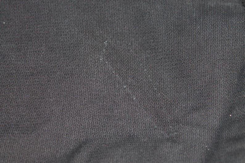 May include: A close-up view of a solid black fabric with a visible woven texture. The material features a consistent dark charcoal to black color throughout, displaying a tight, uniform weave pattern with subtle light-colored lint particles.
