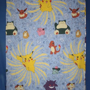 May include: Light blue Pokémon-themed fabric featuring various Pokémon characters like Pikachu, Charmander, and Snorlax. The design includes yellow lightning bolt patterns and the Pokémon logo at the bottom. The fabric is likely for bedding or crafting.