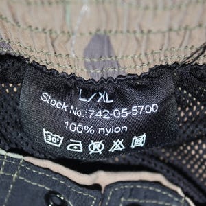 May include: A black interior garment label with white text reading L/XL, Stock No.: 742-05-5700, and 100% nylon. The label includes five laundry care symbols and is attached to a camouflage patterned garment with a black mesh lining and stitching.