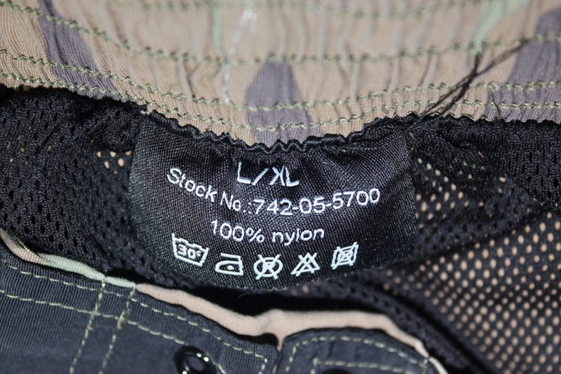 May include: A black interior garment label with white text reading L/XL, Stock No.: 742-05-5700, and 100% nylon. The label includes five laundry care symbols and is attached to a camouflage patterned garment with a black mesh lining and stitching.