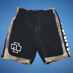 May include: Black board shorts featuring a white Rammstein logo on the lower left leg. The shorts have camouflage side panels and a camouflage hem. The word Rammstein is printed in large white capital letters vertically along the right side panel.
