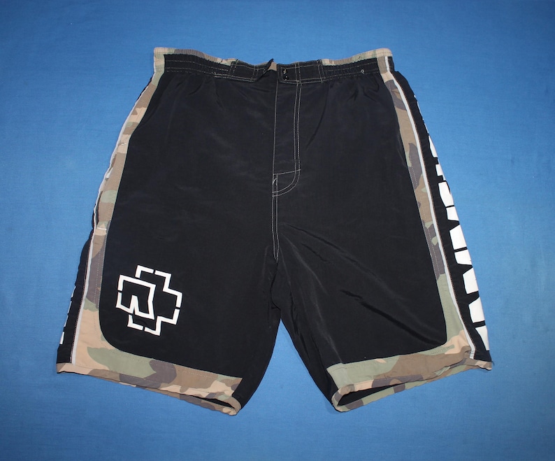 May include: Black board shorts featuring a white Rammstein logo on the lower left leg. The shorts have camouflage side panels and a camouflage hem. The word Rammstein is printed in large white capital letters vertically along the right side panel.