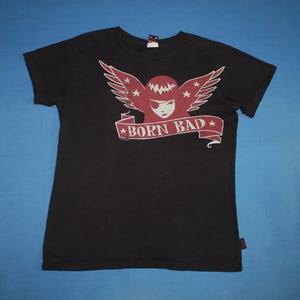May include: Black t-shirt with a red and white graphic. The graphic features a winged figure with an eye patch and the text "BORN BAD" on a banner. The shirt has short sleeves and a crew neck.