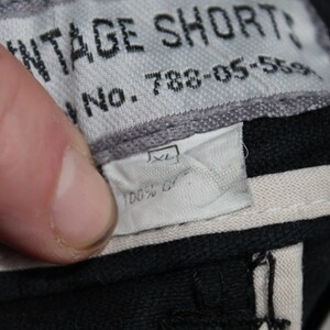 May include: A black garment label with white text reading VINTAGE SHORTS and No. 788-05-5591. Below this, a smaller white tag indicates size XL and 100% cotton. The labels are stitched onto a black fabric base with light-colored interior trim.