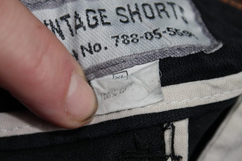 May include: A black garment label with white text reading VINTAGE SHORTS and No. 788-05-5591. Below this, a smaller white tag indicates size XL and 100% cotton. The labels are stitched onto a black fabric base with light-colored interior trim.