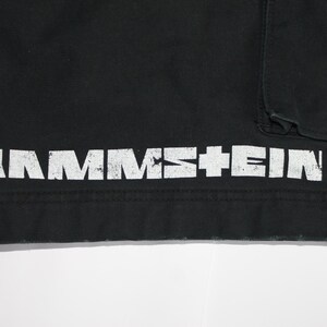 May include: A black garment featuring the white distressed text RAMMSTEIN printed horizontally along the bottom hem. The fabric has a visible seam above the text and a portion of a pocket is visible in the upper right corner of the frame.