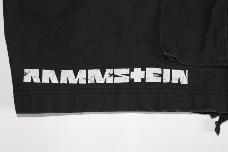 May include: A black garment featuring the white distressed text RAMMSTEIN printed horizontally along the bottom hem. The fabric has a visible seam above the text and a portion of a pocket is visible in the upper right corner of the frame.