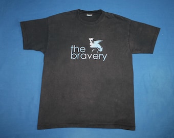 May include: A dark gray t-shirt with the words "the bravery" in light blue, along with a stylized graphic of a face and bird. The shirt is made of a soft material and has short sleeves.